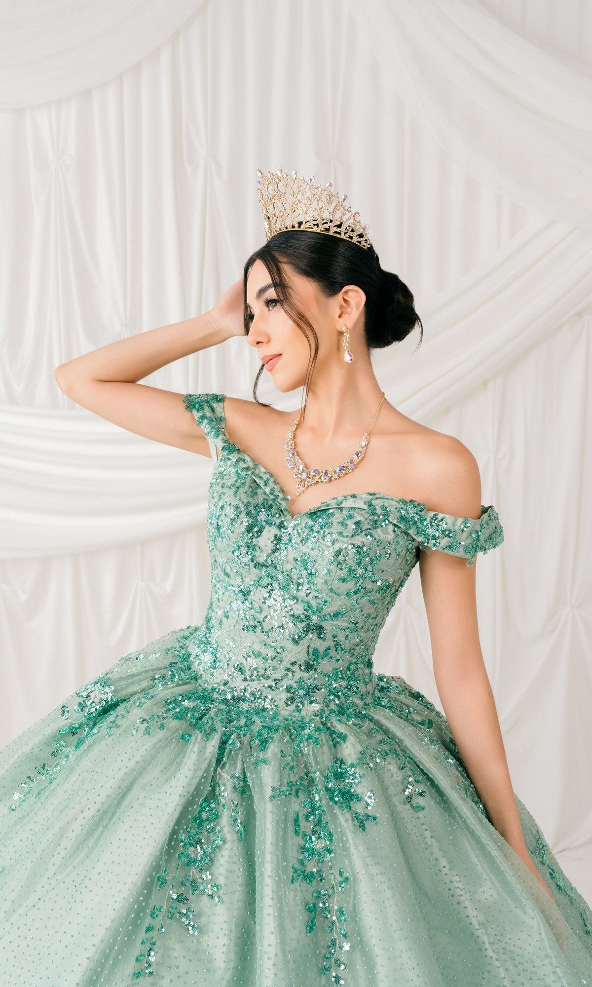 A woman poses against white draped fabric wearing the Quinceanera Ball Gown 8060J—an off-the-shoulder green sequin dress with a lace-up back, styled with a tiara, necklace, and earrings.